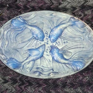 May include: Oval-shaped, light blue glass dish featuring four stylized fish swimming in a circular pattern. The fish have detailed fins and tails, with water and bubbles adding to the aquatic theme. A decorative piece.