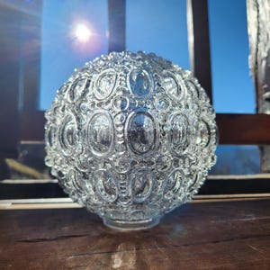 May include: A clear glass globe with an intricate, textured pattern of oval and circular shapes. The orb-shaped object is sitting on a wooden surface, with a bright sun and blue sky in the background. The glass has a vintage, decorative appearance.
