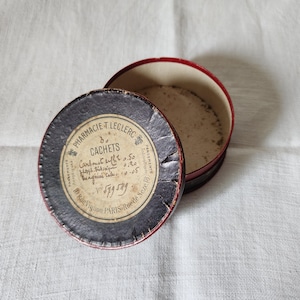 Può includere: Un contenitore rotondo antico aperto, con bordo rosso e coperchio circolare scuro. Il coperchio presenta un'etichetta con testo francese, tra cui "Pharmacie T. Leclerc" e "Cachets". Il contenitore è su una superficie bianca strutturata.