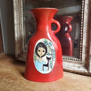 May include: Red ceramic vase with a handle, featuring a painted design of a woman with a cat in a green and white oval frame.