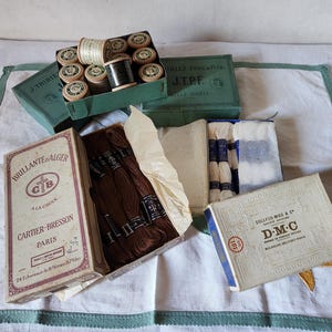May include: A selection of vintage sewing items, including thread spools, embroidery floss, and boxes from brands such as J. Thiriez, Cartier-Bresson, and D.M.C. The items are displayed on a white surface with a green border.