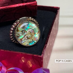 May include: A gold-toned phone ring holder with a round, iridescent abalone shell design. The ring is set in a beaded frame and rests inside a red gift box. The text "POP N CASES" is visible.