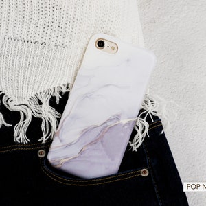 May include: A white marble phone case with a subtle purple and gold swirl design. The case is in a blue jean pocket.