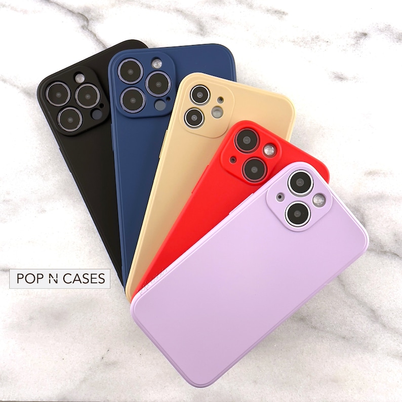 May include: A set of six phone cases in various colours, including black, blue, beige, red, and purple. The cases are designed for a phone with a triple camera lens and feature a raised camera bump for protection. The cases are arranged in a fan shape on a white marble surface. The text "POP N CASES" is visible in the bottom left corner of the image.