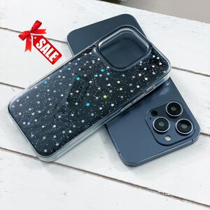 May include: A clear phone case with a black background and a pattern of small, sparkling stars. The case is on top of a dark blue phone. A red bow with the word "SALE" is in the upper left corner.