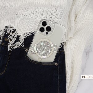 May include: A white phone case with a sparkly, iridescent design is in the front pocket of a pair of blue jeans. The phone case has a clear back and a white ring on the back. The jeans are tucked into a white sweater with a frayed hem. The text "POP N CASES" is visible in the bottom right corner of the image.