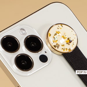 May include: A white iPhone with a gold and white phone grip with a black strap. The grip has gold flecks embedded in the white surface. The strap has the text "POP N CASES" printed on it.