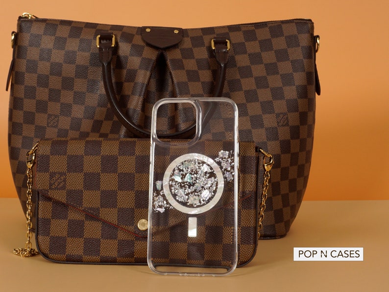 May include: A clear phone case with a silver glitter design and a white circle in the center. The case is sitting on top of a brown and white checkered purse. The text "POP N CASES" is visible in the bottom right corner of the image.