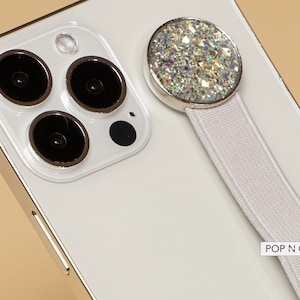 May include: White iPhone with a white Pop N Cases phone grip with a sparkly silver top. The phone has a triple lens camera system.