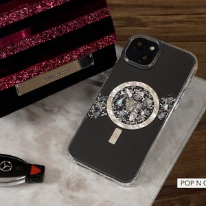 May include: A black iPhone case with a silver and white circular design featuring a variety of small, iridescent pieces. The case is clear on the edges and has a camera cutout. The text "POP N CASES" is visible in the bottom right corner of the image.