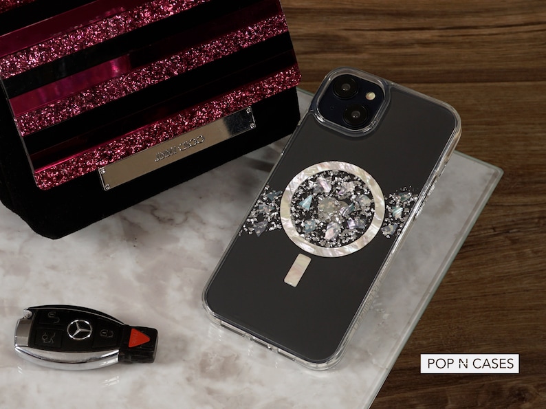 May include: A black iPhone case with a silver and white circular design featuring a variety of small, iridescent pieces. The case is clear on the edges and has a camera cutout. The text "POP N CASES" is visible in the bottom right corner of the image.