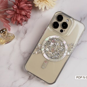 May include: A clear phone case with a white and silver glitter design. The case has a circular design with a pearl-like center. The case is for an iPhone with a triple camera lens. The text "POP N CASES" is visible in the bottom right corner of the image.