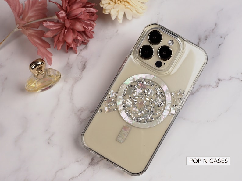 May include: A clear phone case with a white and silver glitter design. The case has a circular design with a pearl-like center. The case is for an iPhone with a triple camera lens. The text "POP N CASES" is visible in the bottom right corner of the image.