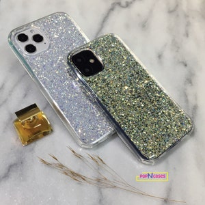 May include: Two clear phone cases with a sparkly glitter design. The case on the left is white with iridescent glitter, and the case on the right is gold with green glitter. The cases are on a white marble surface.