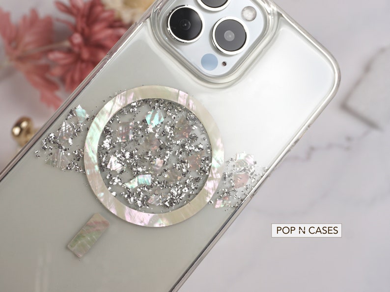 May include: A clear phone case with a silver glitter and iridescent shell design. The case has a raised circle with a shell design and silver glitter inside. The text "POP N CASES" is visible in the bottom right corner of the image.