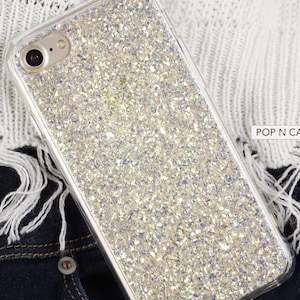 Könnte beinhalten: Eine transparente Handyhülle mit einem glitzernden silbernen Glitzerdesign. Die Hülle ist aus strapazierfähigem Kunststoff gefertigt und kompatibel mit den meisten Smartphones. Die Hülle wird auf einem weißen gestrickten Stoffhintergrund gezeigt.