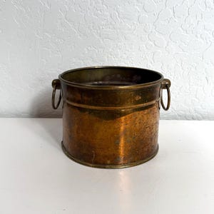 Puede incluir: Una olla cilíndrica de cobre con un acabado metálico desgastado y una pátina oscura. El recipiente cuenta con dos pequeñas asas circulares de latón unidas a los lados opuestos cerca del borde superior, ofreciendo un diseño sencillo.