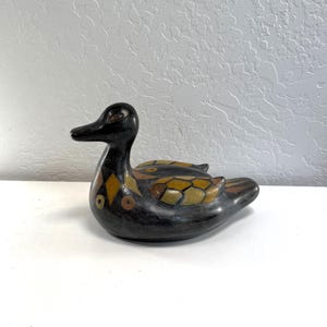 以下が含まれることがあります： 黒い陶器製のカモの置物で、滑らかで暗い仕上げが施されています。胸と翼には黄色、オレンジ、茶色の幾何学模様が装飾されています。カモは頭を横に向けた休息の姿勢で表現されています。