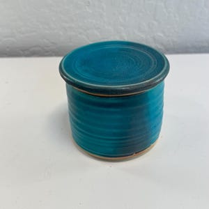 May include: A cylindrical ceramic jar with a matching lid, featuring a vibrant teal blue glaze with subtle horizontal ribbed textures along the body and a smooth, slightly darker rim on the lid, showcasing a glossy finish throughout the piece.