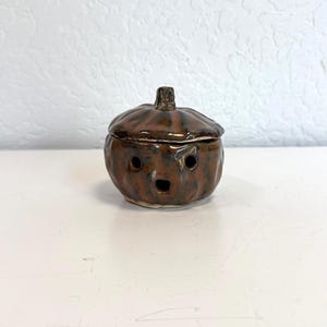 May include: A ceramic pumpkin-shaped container with a lid. The container is brown with darker brown stripes and a glossy finish. The pumpkin has a surprised expression with two eye holes and a mouth hole.