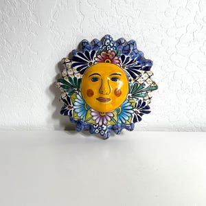 Puede incluir: Un colgante de pared con cara de sol de cerámica pintada a mano. El sol tiene una cara amarilla con mejillas rosadas, ojos azules y una expresión sonriente. El sol está rodeado por un borde azul, verde y con motivos florales. Un artículo de decoración único.
