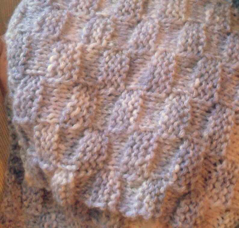 K&ouml;nnte beinhalten: Nahaufnahme eines gestrickten Stoffes mit einem strukturierten Muster. Der Stoff ist helllila mit einer dezenten wei&szlig;en Variation.