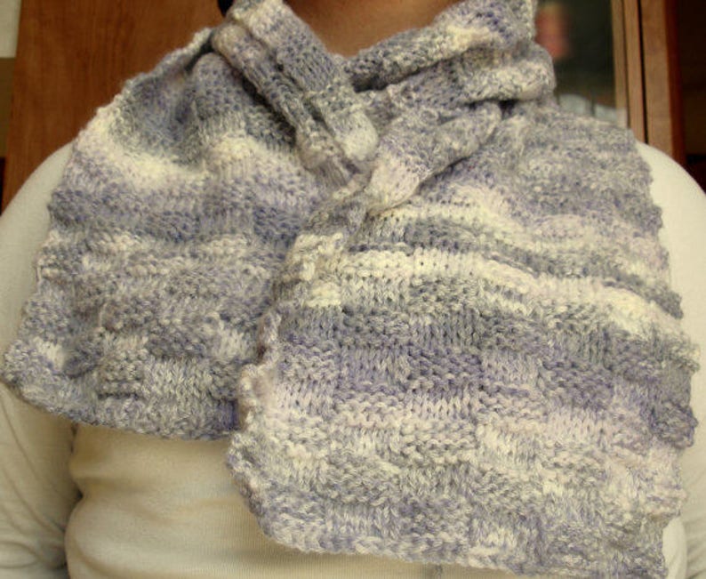 K&ouml;nnte beinhalten: Ein weicher, gestrickter Schal in Wei&szlig;-, Grau- und Lilat&ouml;nen. Der Schal ist aus einem dicken Garn gefertigt und hat eine strukturierte, gewebte Optik.