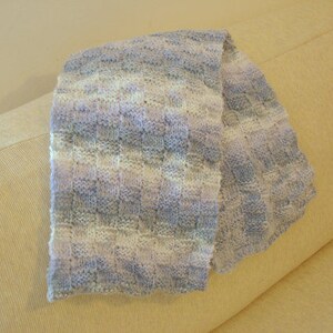 K&ouml;nnte beinhalten: Ein weicher, gestrickter Schal in Wei&szlig;-, Hellblau- und Lavendelt&ouml;nen. Der Schal ist gefaltet und &uuml;ber ein hellbeiges Sofa gelegt.