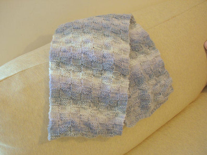 K&ouml;nnte beinhalten: Ein weicher, gestrickter Schal in Wei&szlig;-, Hellblau- und Lavendelt&ouml;nen. Der Schal ist gefaltet und &uuml;ber ein hellbeiges Sofa gelegt.