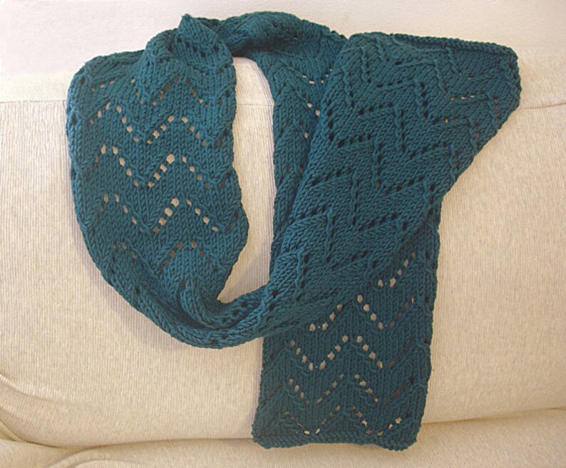 May include: A teal blue knitted scarf with a lacy pattern. The scarf is folded in half and draped over a light beige surface.