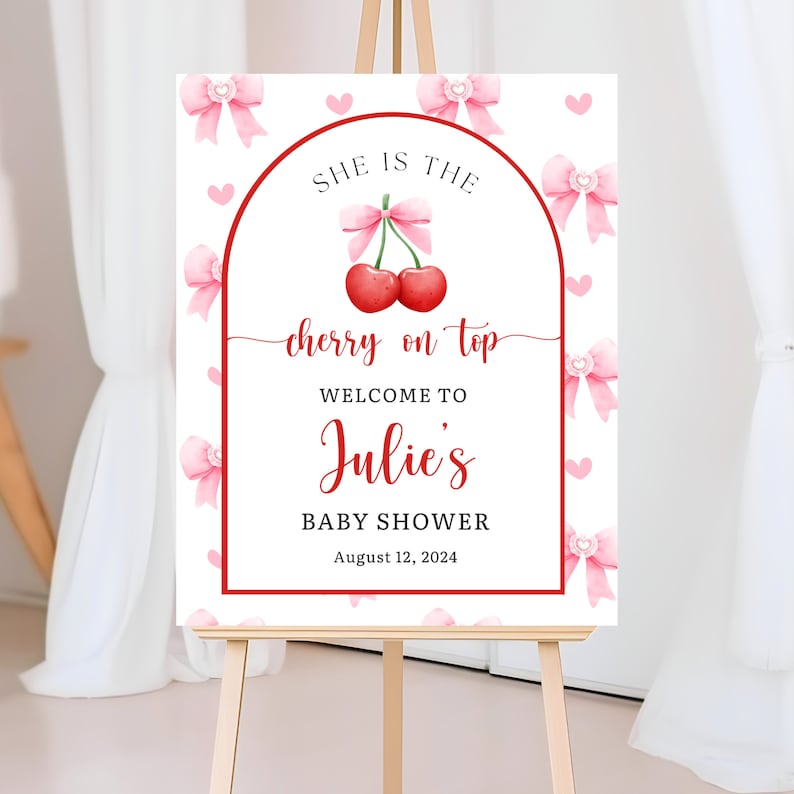 May include: A white sign on an easel with a red border and text. The sign reads "SHE IS THE cherry on top WELCOME TO Julie's BABY SHOWER August 12, 2024." Pink bows and hearts decorate the background. A pair of cherries is in the center.