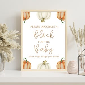 May include: A framed sign with a light wood border. The sign has the text "PLEASE DECORATE A Block FOR THE Baby" in a stylish font. Watercolor pumpkins in orange and white decorate the corners and center. The text "Don't forget to sign your name!" is at the bottom.
