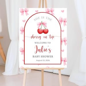 May include: A white sign on an easel with a red border and text. The sign reads "She is the cherry on top, Welcome to Julie's Baby Shower, August 12, 2024." Pink bows and hearts decorate the background.