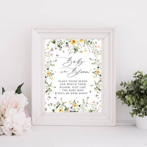 Op de afbeelding: Een wit ingelijst print met een bloemenontwerp en de tekst "Baby in Bloom" met de boodschap "Plant deze zaden en bekijk hoe ze bloeien, net als de baby die er binnenkort zal zijn!"