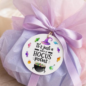 May include: A round white gift tag tied with a lavender satin ribbon to a lavender tissue paper bundle. The tag features the black text It's just a bunch of HOCUS POCUS, a purple witch hat, a black cauldron with colorful candies, and small candy icons.