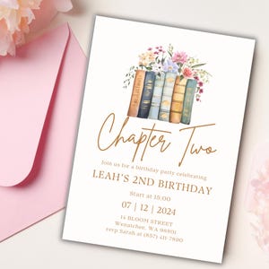Puede incluir: Una tarjeta de invitación blanca con un borde y texto dorados. La tarjeta presenta una ilustración de acuarela de una pila de libros con flores en la parte superior. El texto dice "Capítulo Dos" y "El 2º cumpleaños de Leah". La fecha y la hora de la fiesta se enumeran a continuación, junto con la dirección y la información de RSVP.