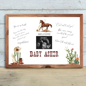 Puede incluir: Un cartel enmarcado en blanco con un marco de madera marrón. El cartel tiene una ilustración de acuarela de un cactus, un sombrero de vaquero y un caballo. El texto en el cartel dice "You are so loved!" "May you dream little baby" "So joyous and new" "Bring a whole world of wonderful" "Blessings to you" "Congratulations!" "Welcome" "Can't wait to meet you!" "We are very excited to meet you!" "Welcome to the world little one!" "BABY ASHER".