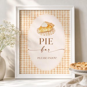 May include: A framed sign with a light orange and white gingham background featuring an oval center. The sign displays a watercolor illustration of a slice of pie with whipped cream, the text PIE bar, and the words PLEASE ENJOY! in dark brown.