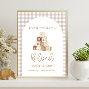 May include: A framed sign with a tan and white gingham pattern border. The sign reads "PLEASE DECORATE A BLOCK FOR THE BABY" with a stack of letter blocks. The text "Don't forget to sign your name!" is at the bottom.