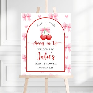 May include: A baby shower welcome sign on an easel. The sign features a red cherry design with the words "She is the cherry on top" and "Welcome to Julie's Baby Shower August 12, 2024". Pink bows and hearts decorate the white background.