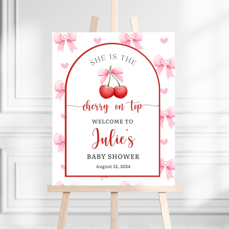 May include: A baby shower welcome sign on an easel. The sign features a red cherry design with the words "She is the cherry on top" and "Welcome to Julie's Baby Shower August 12, 2024". Pink bows and hearts decorate the white background.