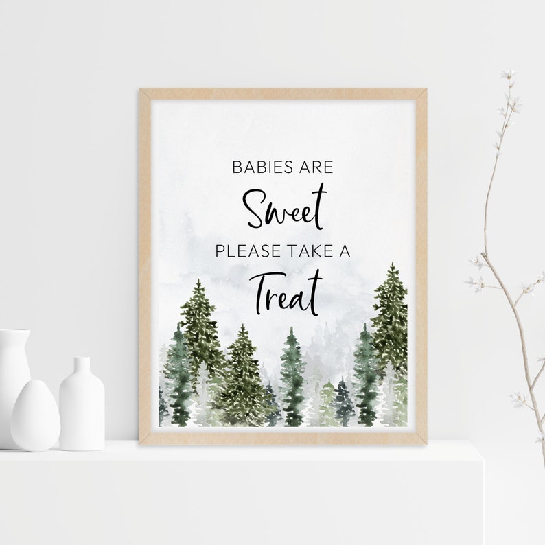 May include: A framed sign with a light wood border featuring a watercolour forest of green pine trees at the bottom and the black text BABIES ARE Sweet PLEASE TAKE A Treat centred on a white background with a soft grey misty effect.