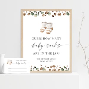May include: A framed baby shower game sign with the text "Guess How Many Baby Socks Are in the Jar?" and a matching card. The sign features watercolor illustrations of baby socks, coffee beans, and greenery.