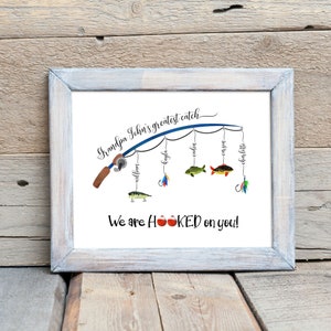 May include: A framed print with a blue fishing rod and the text "Grandpa John's greatest catch".  Five fish are hanging from the line with names written on them: William, Layla, Camden, Carson, Charlotte. The text "We are HOOKED on you!" is below the fishing rod.
