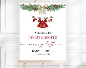 May include: A white sign on an easel with a Christmas-themed baby shower design. The sign features a red dress, hat, and shoes hanging on a clothesline, with text that reads "Welcome to Ashley & Scott's merry little baby shower December 11, 2025."