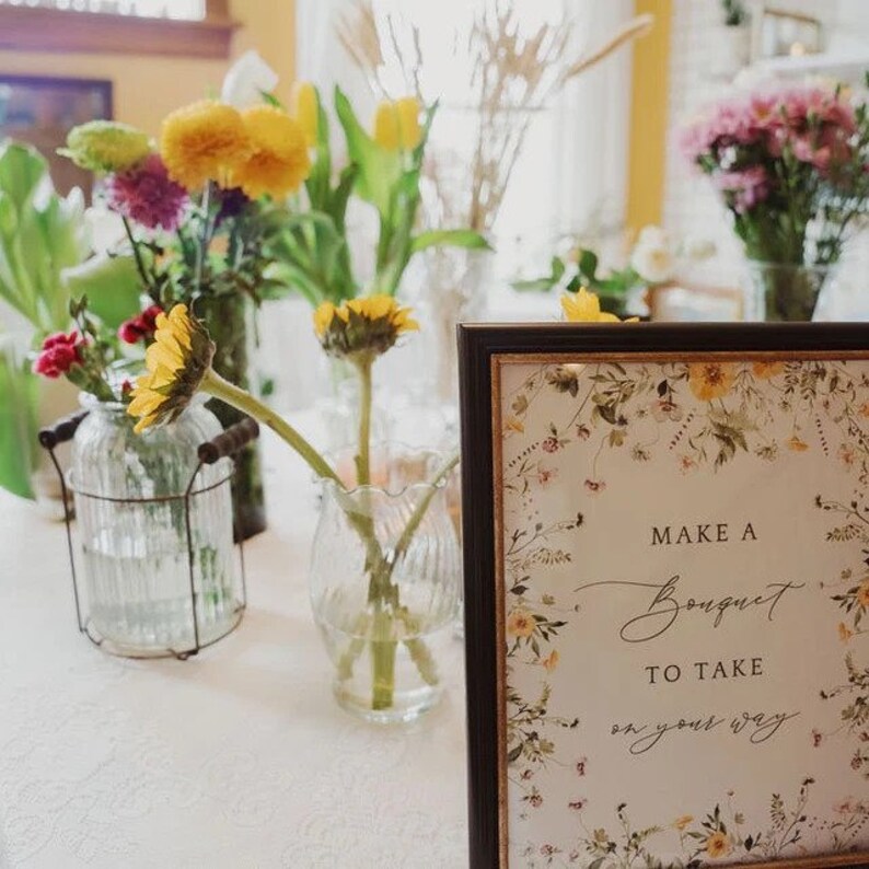 Op de afbeelding: Een ingelijst bord met een bloemenontwerp en de tekst "Make a Bouquet to Take on your way". Het bord staat op een tafel met verschillende vazen met bloemen, waaronder zonnebloemen, gele rozen en roze bloemen.