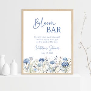 Op de afbeelding: Een wit bord met blauwe tekst en bloemillustraties. Het bord staat "Bloom Bar" en "Maak je eigen boeket om mee naar huis te nemen aan het einde van de dag!" Het staat ook "Victoria's Shower" en "May 11, 2025" erop.