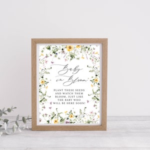Op de afbeelding: Een ingelijst afdruk met een bloemenontwerp en de tekst "Baby in Bloom." De tekst onder het bloemenontwerp luidt: "Plant deze zaden en bekijk hoe ze bloeien, net als de baby die er binnenkort zal zijn!"