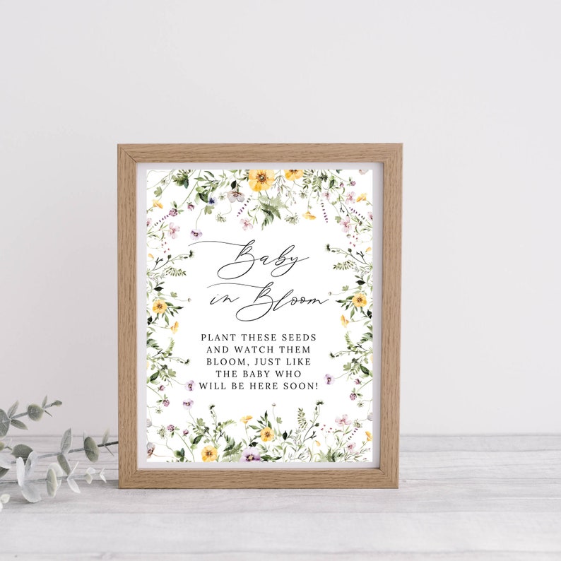 Op de afbeelding: Een ingelijst afdruk met een bloemenontwerp en de tekst "Baby in Bloom." De tekst onder het bloemenontwerp luidt: "Plant deze zaden en bekijk hoe ze bloeien, net als de baby die er binnenkort zal zijn!"