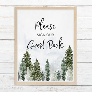 May include: A rectangular sign in a light wood frame featuring the black text Please SIGN OUR Guest Book centred above a watercolour illustration of a dense forest with various shades of green pine trees against a misty white background.